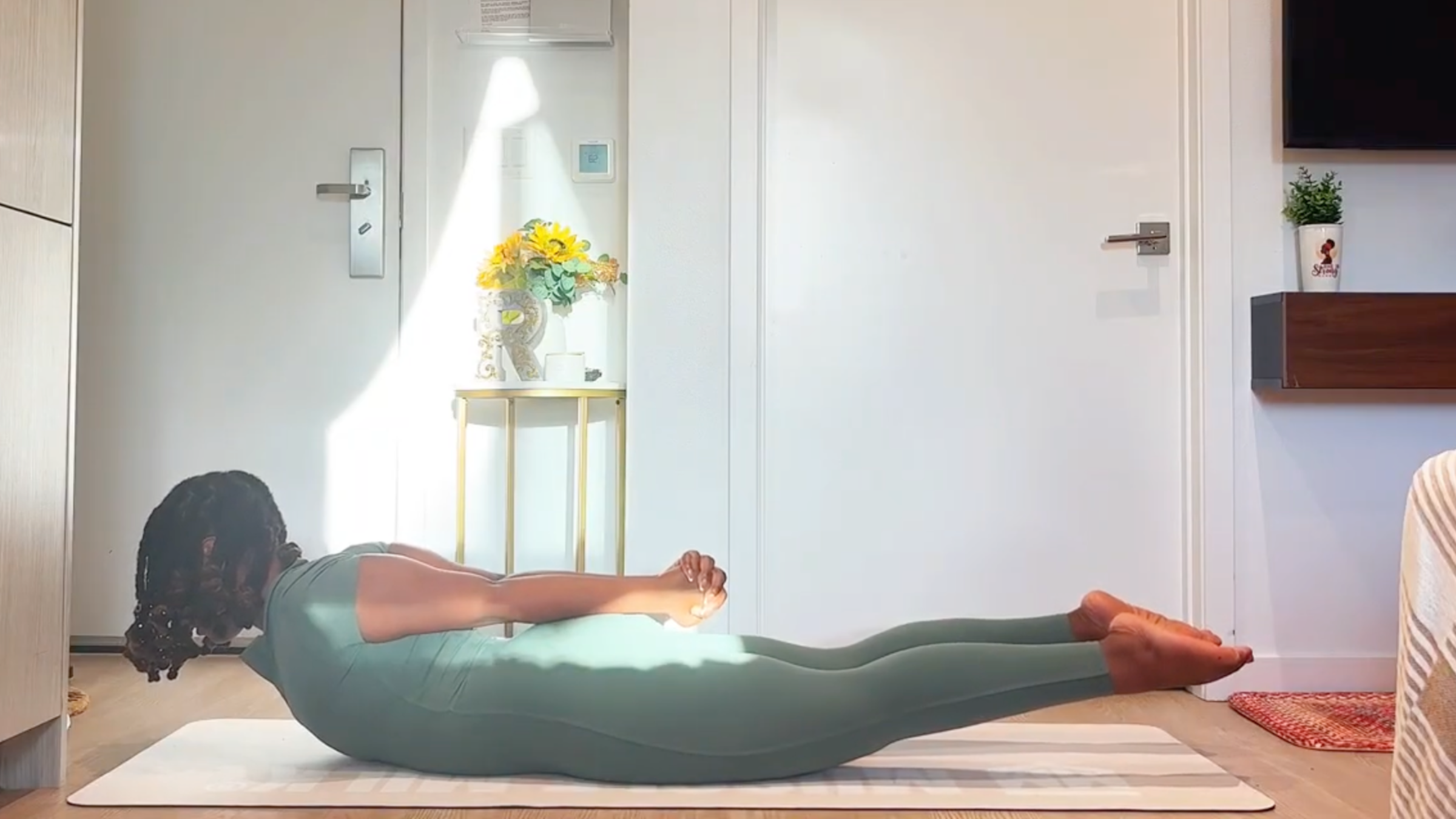 Ramoni Overton lying prone on a yoga mat with hands clasped behind back, lifting chest and legs into Locust pose.